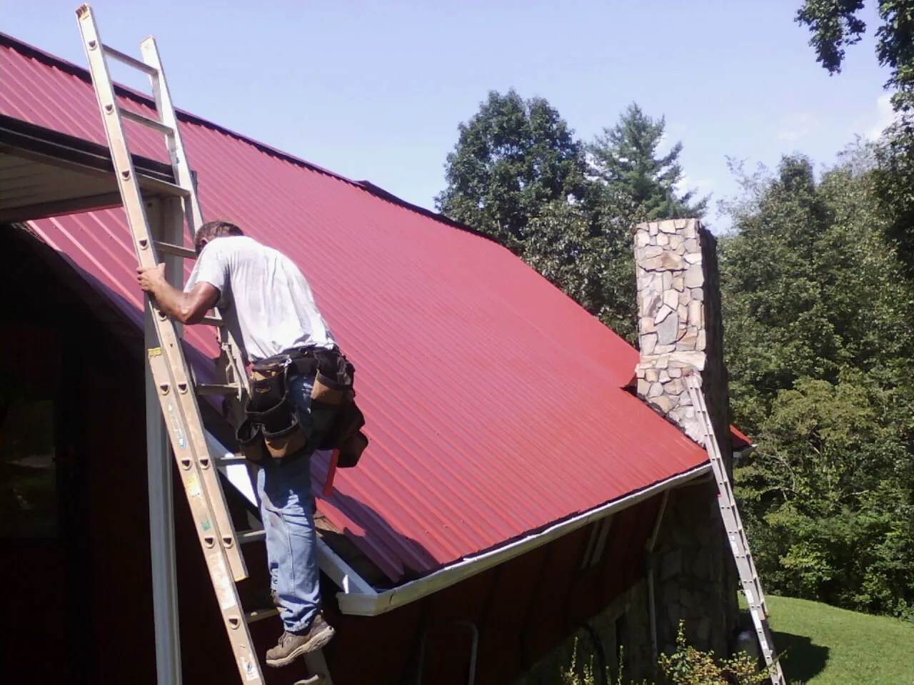 Roofer on a ladder beside a red metal roof and stone chimney — Soffit & Fascia Repair by Roofing Brothers in Heppner